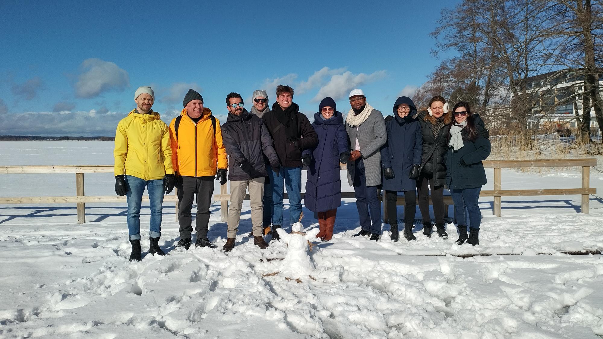 SkillsRec -hankkeen kansainväliset vieraat ja lumiukko seisovat aurinkoisen ja lumisen järven rannalla.